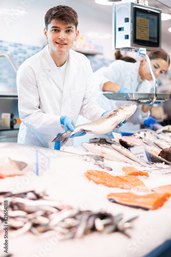 Proficient shop assistant in work clothes holding in hands seabass in fish shop with large stock