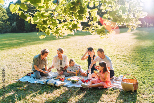 Billede på lærred Big family under Linden tree on the picnic blanket on the in city park green grass