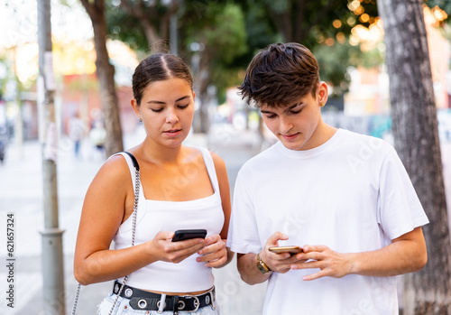 Young man and woman using smartphones outdoors while walking through city street.