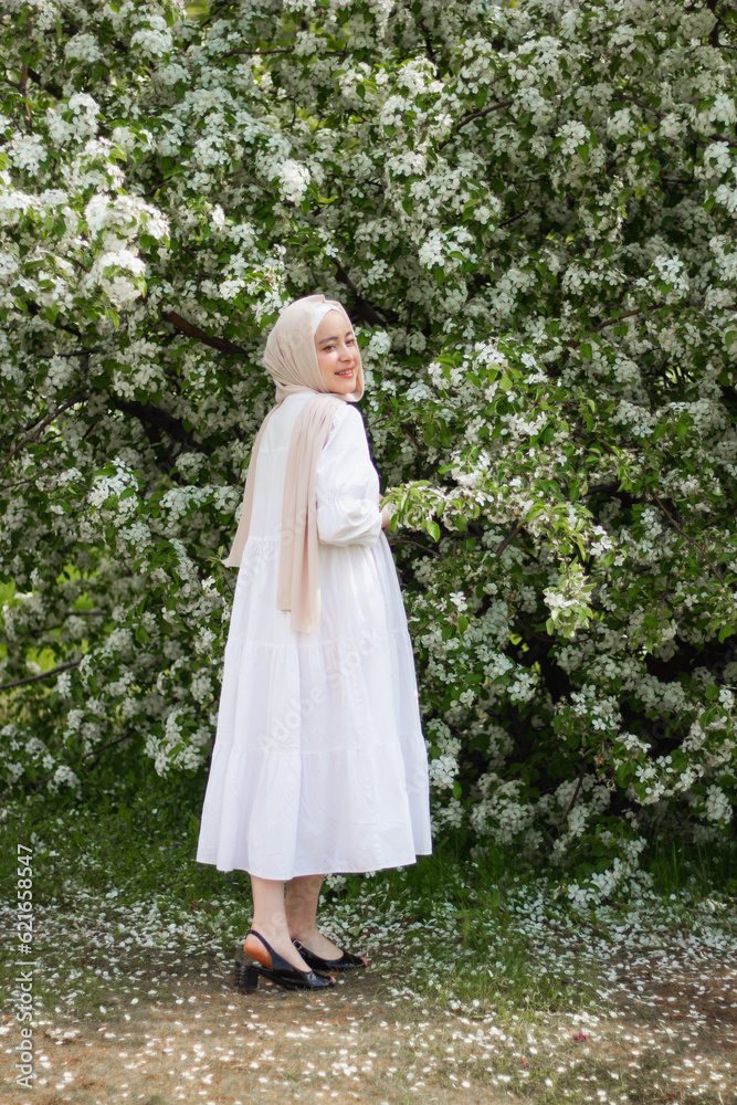 A young Muslim woman in a white dress in front of a flowering tree. In full growth. Overall plan. Blooming apple tree. Pretty modest Muslim woman. European Muslim. tenderness. beauty.