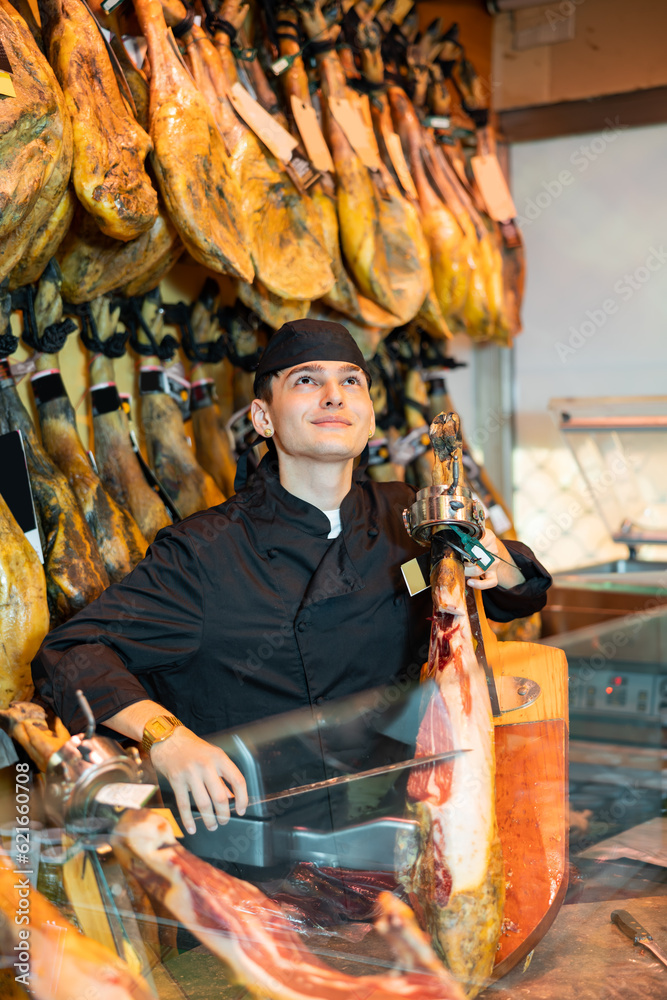 Positive young man working in jamoneria, standing at counter and cutting ham with special knife.