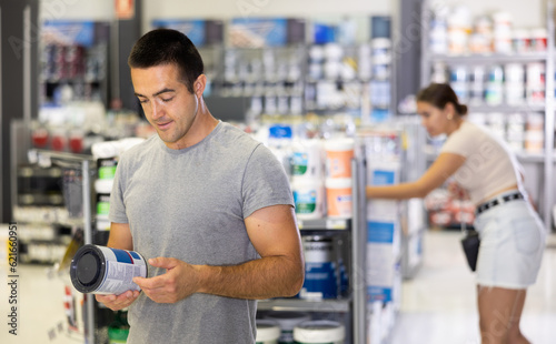 In store, male buyer doubts choice and examines product, paint jar studies detailed information on packaging, label. Repair and construction items, paint and varnish products, accessories, supermarke.