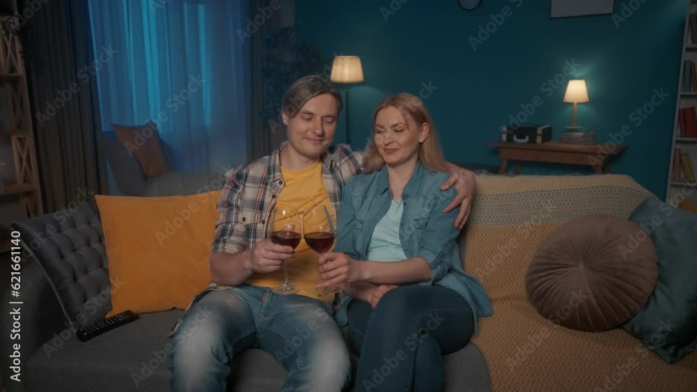 A young couple sits hugging on the couch with glasses of red wine and watching TV. A man and a woman are watching a movie, a wedding video, clinking and drinking wine.