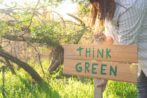 young woman with a hat in an environment of trees and sun with a wooden sign that says think green to protect the natural environment and take care of the environment