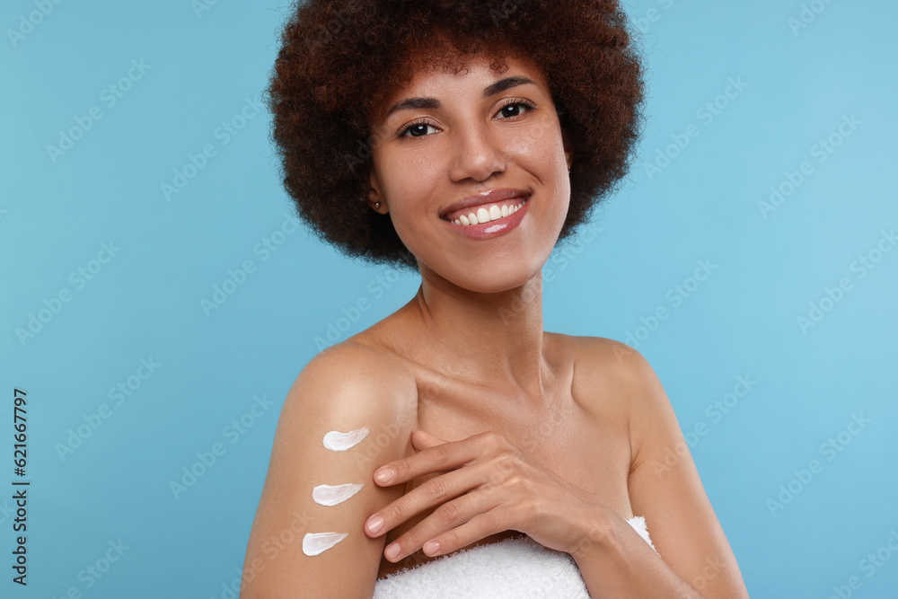 Beautiful young woman applying body cream onto arm on light blue background Beautiful young woman applying body cream onto arm on light blue background