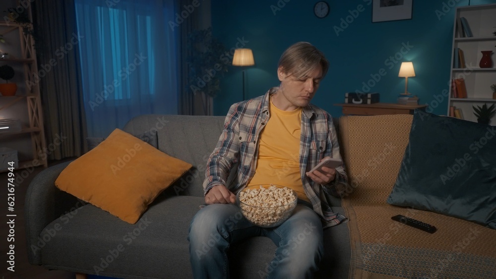 A man is sitting on the couch with a plate of popcorn in front of the TV. A man not interested in a movie sits with a phone in his hands, typing a message, checking email, social network. Boring film. A man is sitting on the couch with a plate of popcorn in front of the TV. A man not interested in a movie sits with a phone in his hands, typing a message, checking email, social network. Boring film.