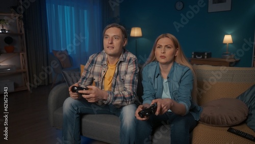 A young couple is playing a video game console with game controllers while sitting on the couch at home in the evening. Man and woman spend time together enjoying the game. Who will win. Game concept.