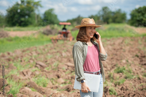 Fotografija Young woman standing on field in sunset while tractor baling in background, Farmer or agrarian with smart phone looking on growing efficiency and crop yield, Smart agriculture and automation concept