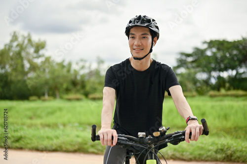 A happy Asian man enjoys listening to music through his earbuds while riding a bike © bongkarn