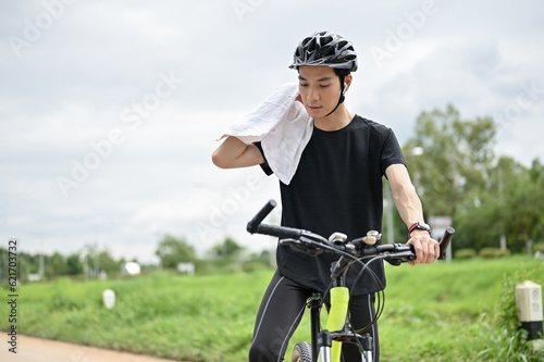 A tired Asian man wiping his neck with a towel while biking along the country roads.