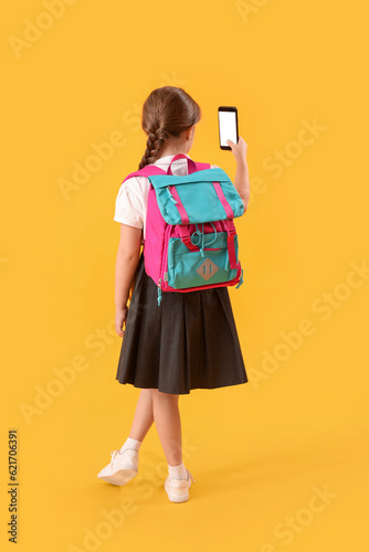 Little schoolgirl with backpack and mobile phone on yellow background, back view