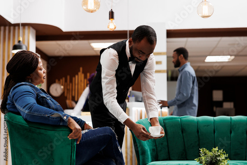 African American guy waiter bringing coffee to guest resting in hotel bar, restaurant worker holding cup of hot beverage serving it to tourist waiting for room in lobby. Hospitality customer service