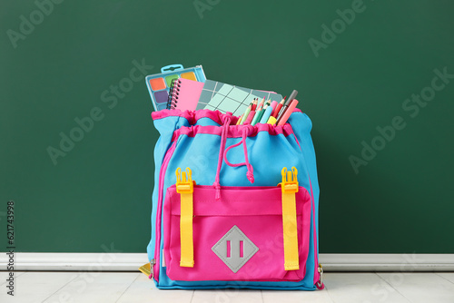 Colorful school backpack with notebooks, watercolors and pencils on white tile table near green chalkboard