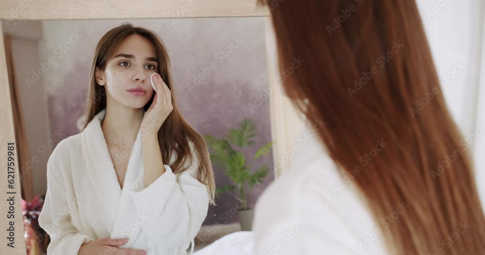 Young brunette woman in a white bathrobe look in the big mirror at home and removing makeup from her face with cotton pad. Skin care. Beautiful woman in a white bathrobe look in the big mirror at home Young brunette woman in a white bathrobe look in the big mirror at home and removing makeup from her face with cotton pad. Skin care. Beautiful woman in a white bathrobe look in the big mirror at home