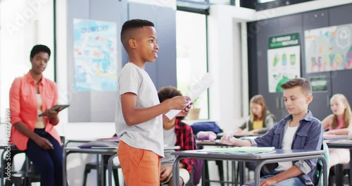 Diverse female teacher and happy schoolchildren at desks reciting in school classroom photo