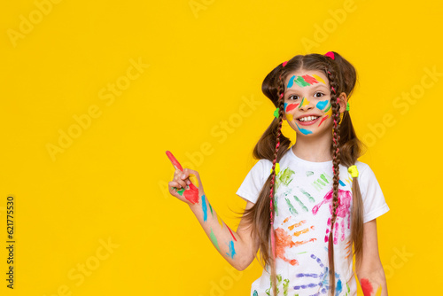 A child smeared with paint. A little girl painted with multicolored paints points to your advertisement. Children's creativity. Yellow isolated background. Copy space. Banner.