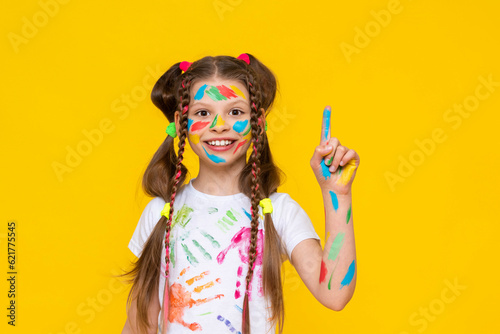 A charming child with painted palms with multicolored paints shows one finger. A little girl happily draws with her hands. Yellow isolated background.