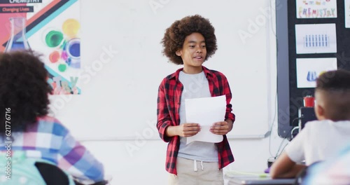 Diverse female teacher and happy schoolchildren at desks reciting in school classroom