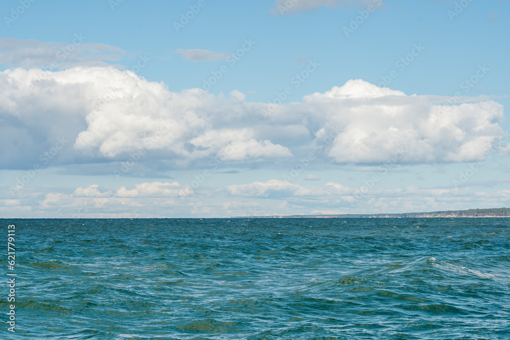 Fototapeta premium View of the sea with the coast in the distance. Calm waves with a blue cloudy sky in warm, summer weather.
