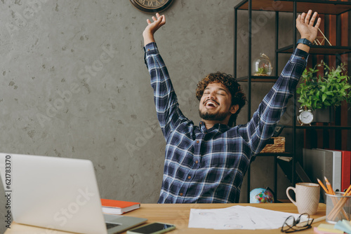 Successful overjoyed employee business Indian man he wears casual blue checkered shirt doing winner gesture celebrate raise up hands finish job sit work at office desk with laptop pc computer indoors.
