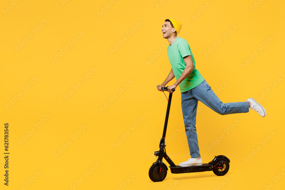Full body sideways young man of African American ethnicity he wearing casual clothes green t-shirt hat riding electric scooter isolated on plain yellow background studio portrait. Lifestyle concept. Full body sideways young man of African American ethnicity he wearing casual clothes green t-shirt hat riding electric scooter isolated on plain yellow background studio portrait. Lifestyle concept.