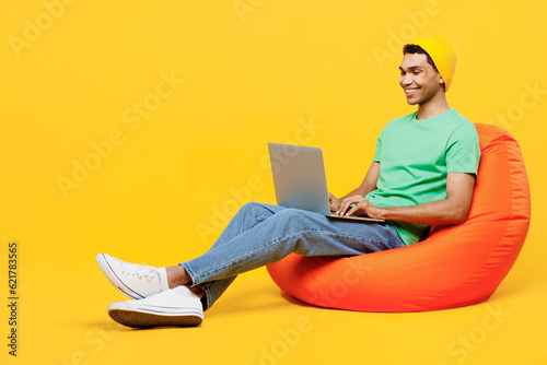 Full body young IT man of African American ethnicity he wears casual clothes green t-shirt hat sit in bag chair hold use work on laptop pc computer isolated on plain yellow background studio portrait.