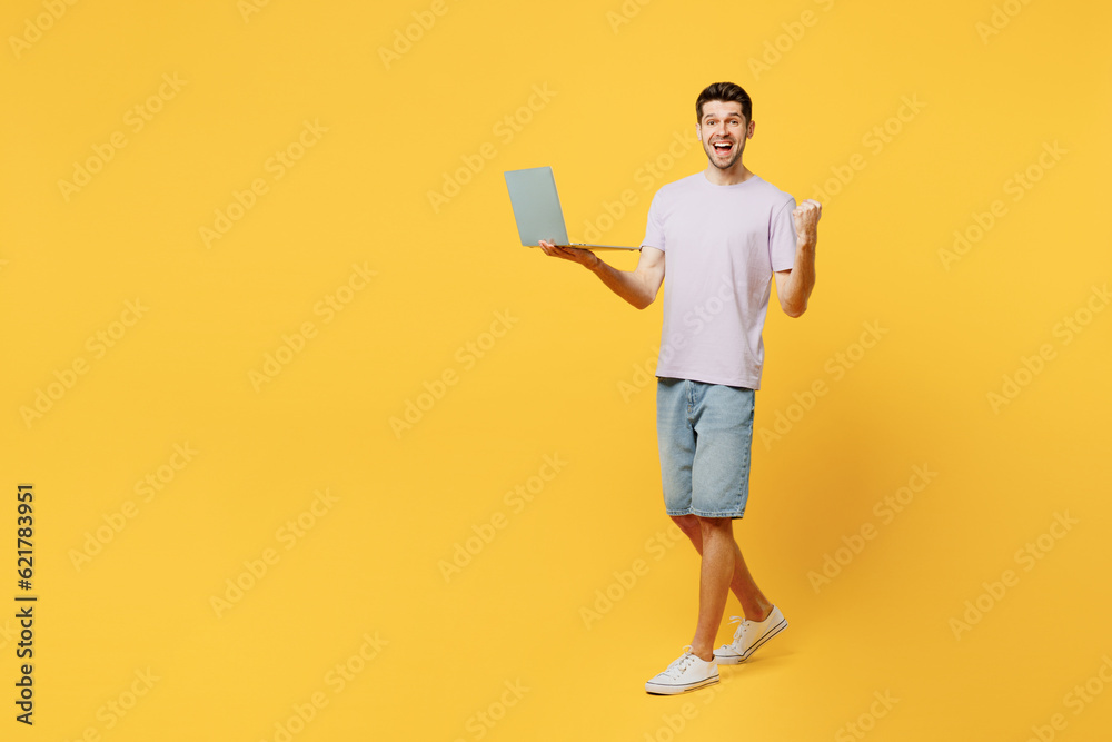 Full body sideways young man he wear light purple t-shirt casual clothes hold use work on laptop pc computer do winner gesture isolated on plain yellow background studio portrait. Lifestyle concept.