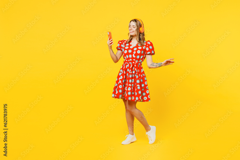 Full body young smiling caucasian woman she wears red dress casual clothes listen to music in headphones use mobile cell phone isolated on plain yellow background studio portrait. Lifestyle concept. Full body young smiling caucasian woman she wears red dress casual clothes listen to music in headphones use mobile cell phone isolated on plain yellow background studio portrait. Lifestyle concept.