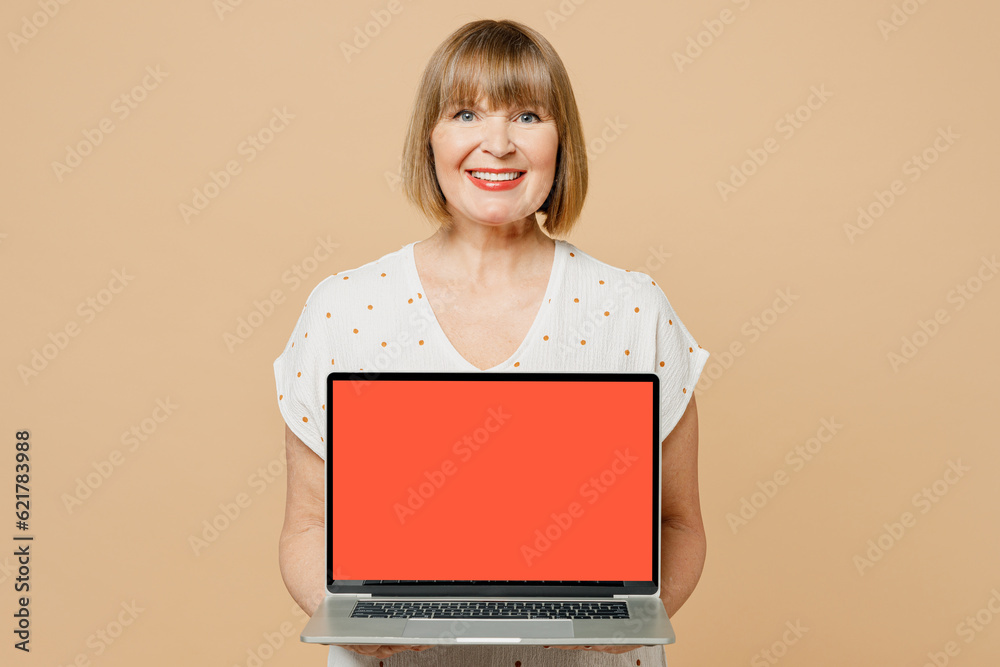 Blonde smiling fun cheerful elderly IT woman 50s years old she wear casual clothes hold use work on laptop pc computer with blank screen workspace area isolated on plain pastel light beige background. Blonde smiling fun cheerful elderly IT woman 50s years old she wear casual clothes hold use work on laptop pc computer with blank screen workspace area isolated on plain pastel light beige background.
