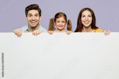 Young smiling parents mom dad with child kid daughter girl 6 years old wearing blue yellow casual clothes hold big white empty poster billboard isolated on plain purple background. Family day concept.