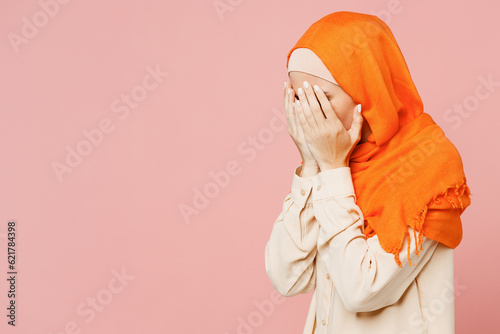 Young sad frustrated arabian asian muslim woman wear orange abaya hijab cover ace with hands crying isolated on plain light pink background studio portrait. Uae middle eastern islam religious concept. photo