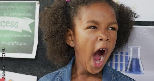 Video portrait of happy, tired biracial schoolgirl yawning in school classroom