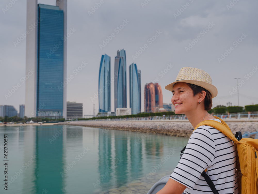 travel to the United Arab Emirates, Happy young asian female traveler with backpack and hat view to cityscape of downtown Abu Dhabi. Vacation and tourist destination concept.
