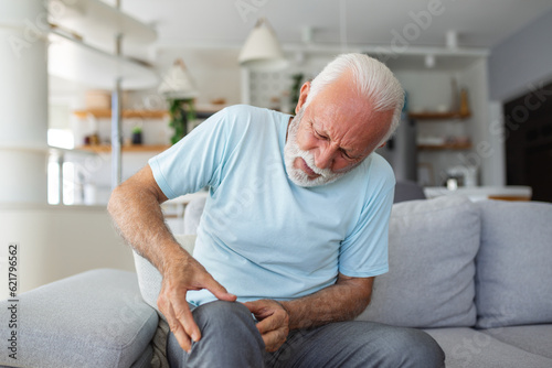Older man sitting on a home couch, holding his leg in pain due to knee joint discomfort, the concept of osteoarthritis, showcasing the challenges faced by individuals with joint-related issues