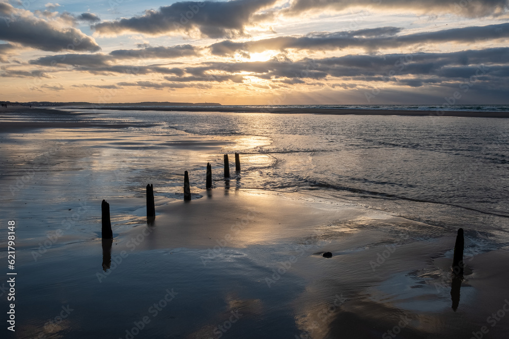 A compelling image showcasing a row of poles on the beach leading into the distance and the waves of the sea. The scene is heightened by a dramatic and colorful sunset sky, painting a striking picture A compelling image showcasing a row of poles on the beach leading into the distance and the waves of the sea. The scene is heightened by a dramatic and colorful sunset sky, painting a striking picture