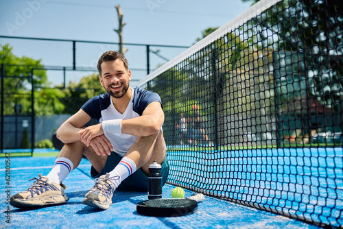 Fototapeta Naklejka Na Ścianę i Meble -  Happy athletic man sitting on paddle tennis court and looking at camera.