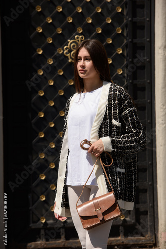 photo of beautiful woman in a white leggings, T-shirt and sweater posing with small brown leather bag