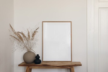 Vertical wooden frame mockup for artwork, photo, print and painting presentation. Black and beige vases with dry grass on wooden bench table. Elegant boho living room interior. White wall, doors.