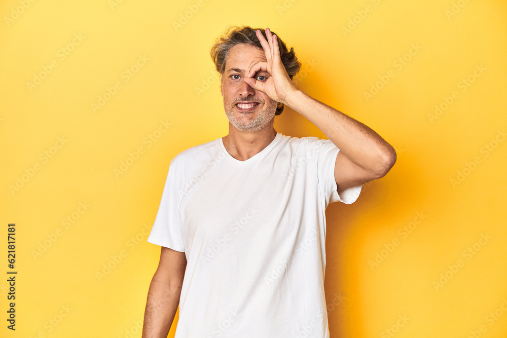 Middle-aged man posing on a yellow backdrop excited keeping ok gesture on eye. Middle-aged man posing on a yellow backdrop excited keeping ok gesture on eye.