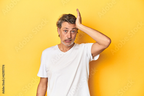 Middle-aged man posing on a yellow backdrop forgetting something, slapping forehead with palm and closing eyes.