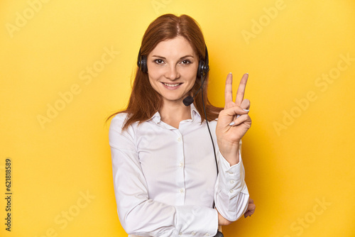 Call center redhead in white shirt, headphones on showing number two with fingers.