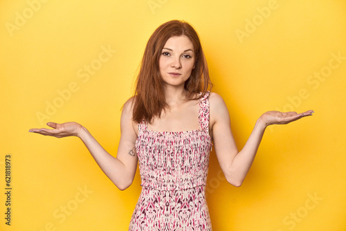 Young redhead in floral dress, studio shot doubting and shrugging shoulders in questioning gesture. © Asier