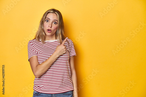 Young blonde Caucasian woman in a red striped t-shirt on a yellow background, pointing to the side