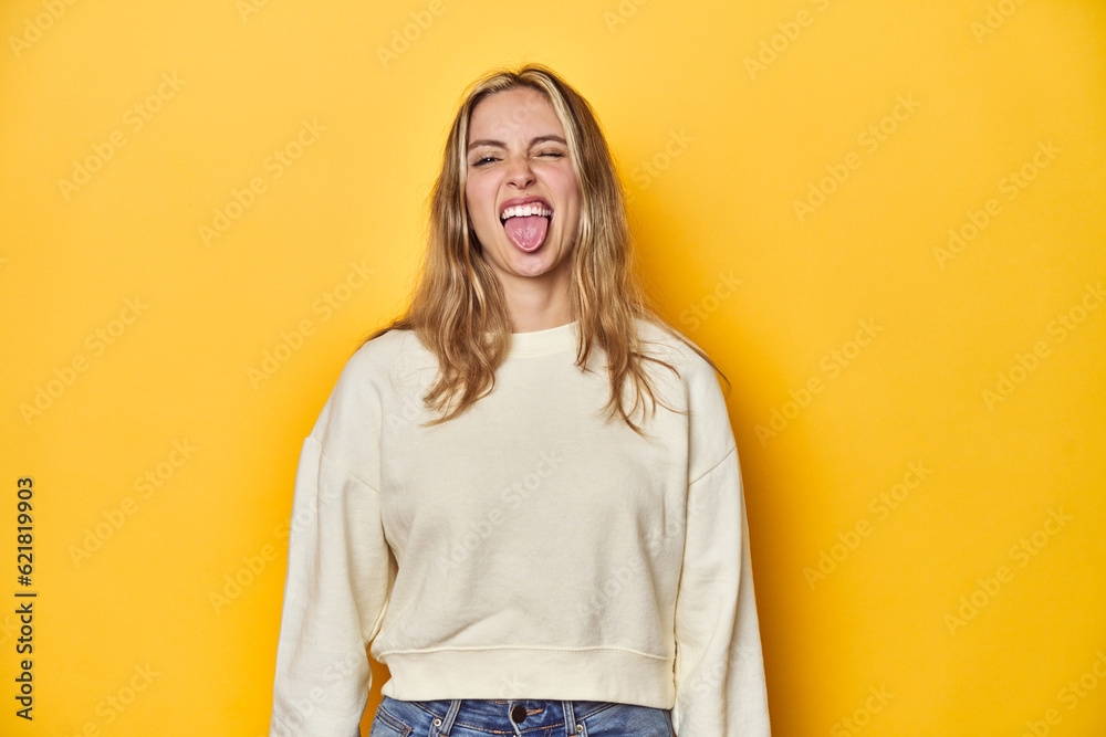 Obraz premium Young blonde Caucasian woman in a white sweatshirt on a yellow studio background, funny and friendly sticking out tongue.