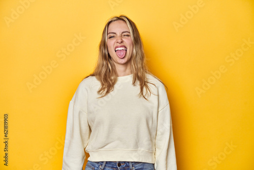 Young blonde Caucasian woman in a white sweatshirt on a yellow studio background, funny and friendly sticking out tongue.