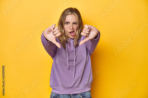 Young blonde Caucasian woman in a violet sweatshirt on a yellow background, showing thumb down and expressing dislike.