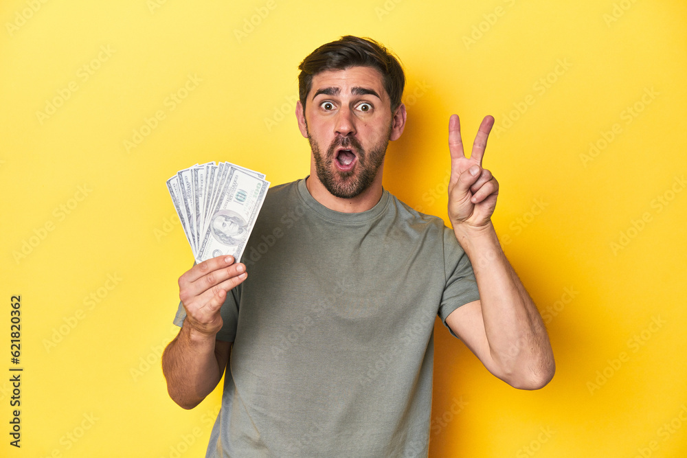 Caucasian man holding dollars, yellow studio shot showing number two with fingers. Caucasian man holding dollars, yellow studio shot showing number two with fingers.