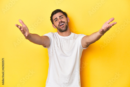 Caucasian man in white t-shirt on yellow studio background feels confident giving a hug to the camera.