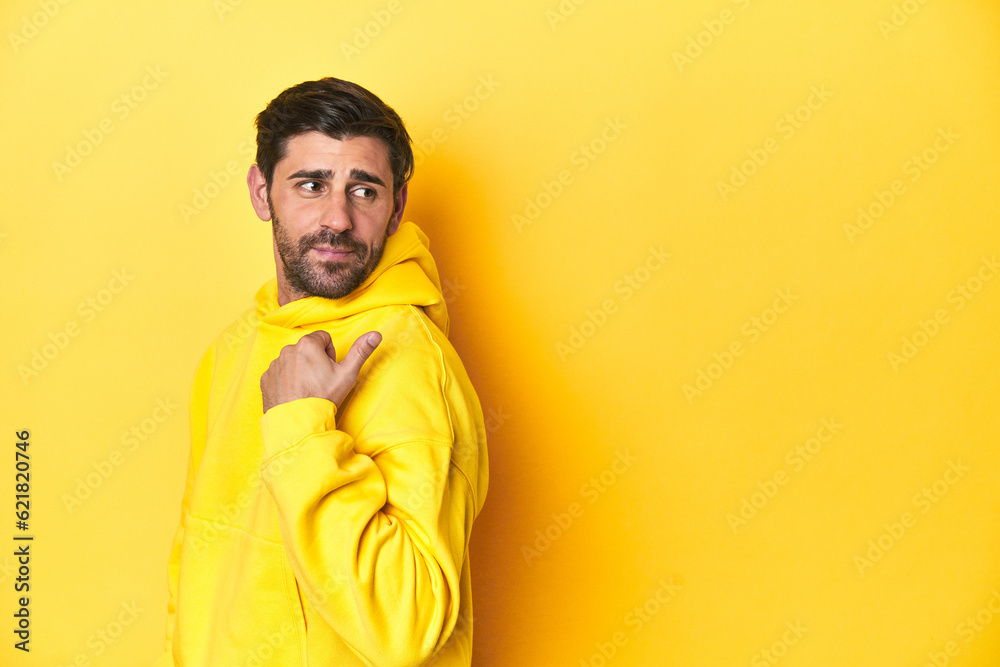 Fototapeta premium Man in yellow hoodie, monochrome studio backdrop points with thumb finger away, laughing and carefree.