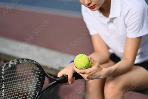 Pretty young sportswoman holding ball racket sitting on the bench at tennis court